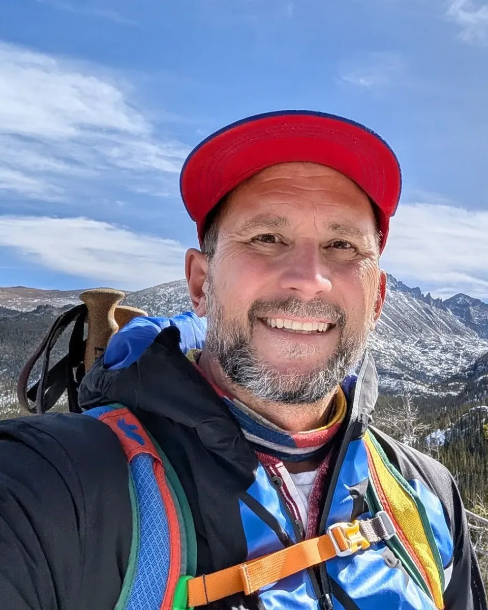 Jim Smith on a mountain hike, smiling, wearing a red cap with snow-covered peaks behind him